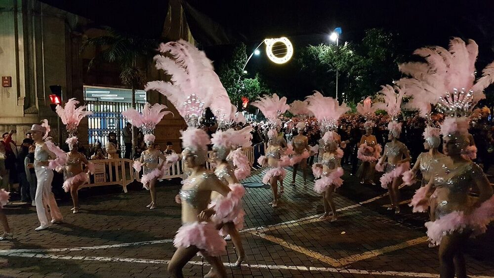 Comparsa de carnaval desfilando de noche con tocados de plumas rosas