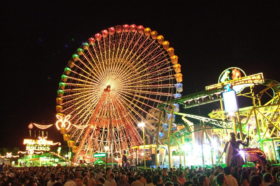 Noria iluminada y atracciones de feria de noche con la multitud en primer plano