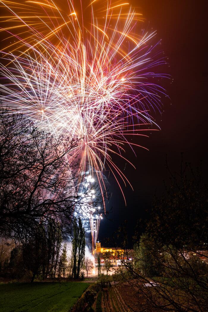 Castillo de fuegos artificiales estallando sobre un pueblo de noche, con la torre de la iglesia iluminada abajo y campos en primer término