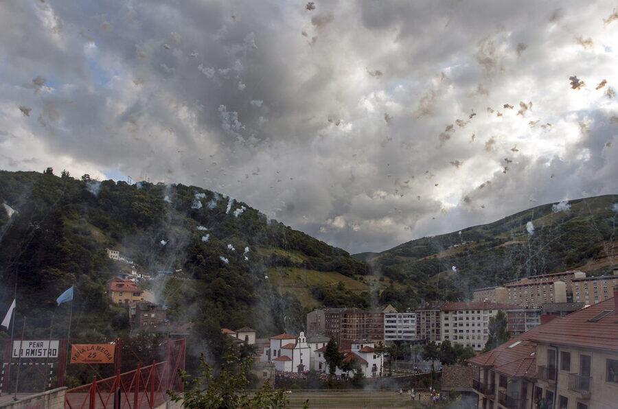 Humo y restos de cohetes diurnos sobre un pueblo encajado en un valle, con pancartas de peñas en primer término