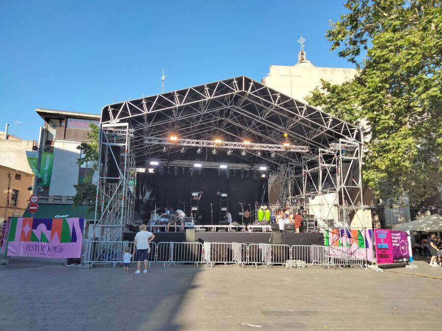 Escenario de estructura metálica montado y vallado en una plaza vacía, con el cartel de la fiesta a un lado