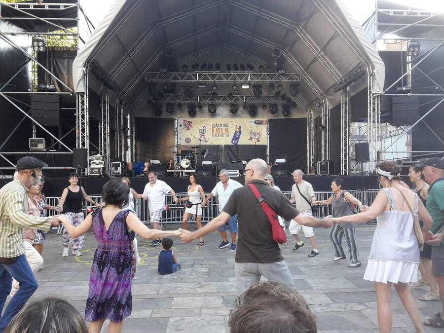 Vecinos de distintas edades bailando en corro cogidos de la mano en una plaza, delante de un escenario pequeño con el cartel de la fiesta