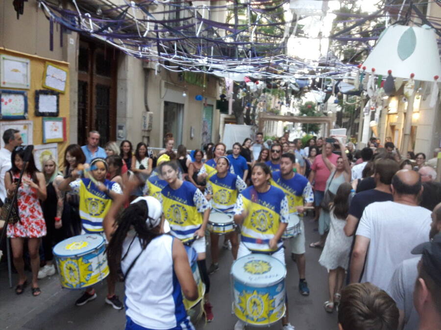 Calle estrecha completamente cubierta de adornos colgantes y abarrotada de gente mientras pasa una batucada con bombos y cajas