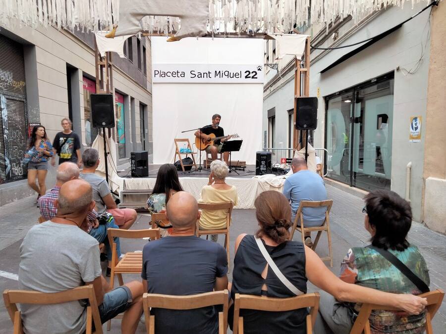 Treinta sillas de madera colocadas en una calle estrecha engalanada, con el público de espaldas y un guitarrista solo sobre un pequeño escenario al fondo