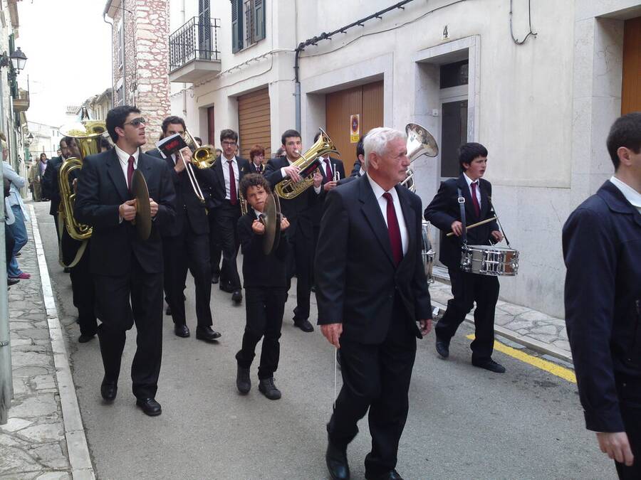 Banda de música municipal recorriendo una calle estrecha, con metales, caja y platillos, encabezada por un hombre mayor de traje oscuro