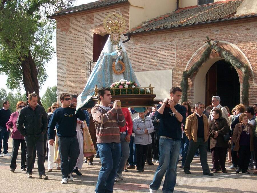 Cuatro vecinos portando sobre los hombros unas andas con la imagen de la patrona a la salida de una ermita de ladrillo, rodeados de público