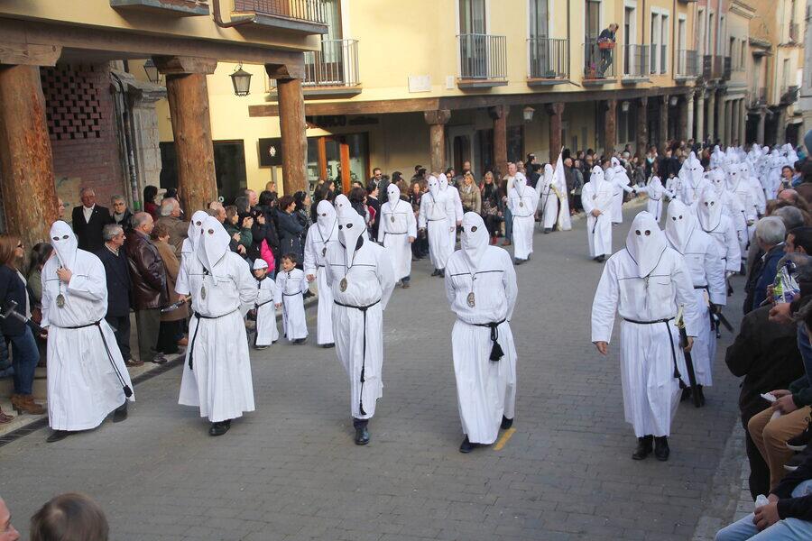 Desfile de cofradías con túnicas blancas por una calle porticada, con el público agolpado a los lados