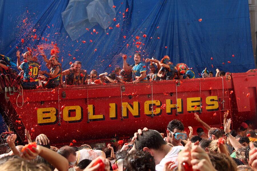 Camión rojo cargado de tomates rodeado de una multitud en una calle estrecha durante la Tomatina de Buñol
